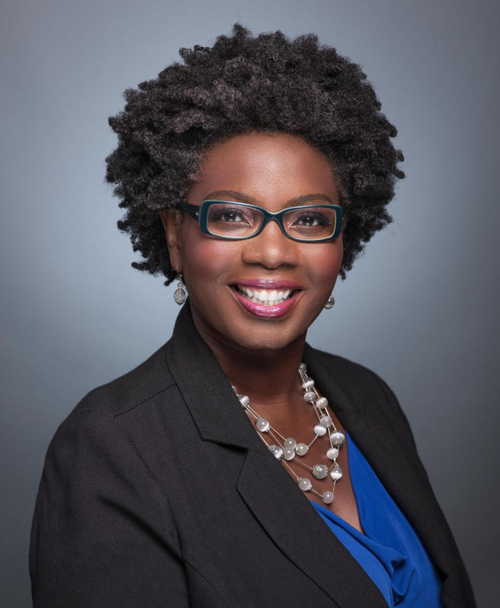 Regina Nunnally Headshot of Regina wearing black rimmed glasses and black suit jacket, around her neck is a silver necklace matching her earrings